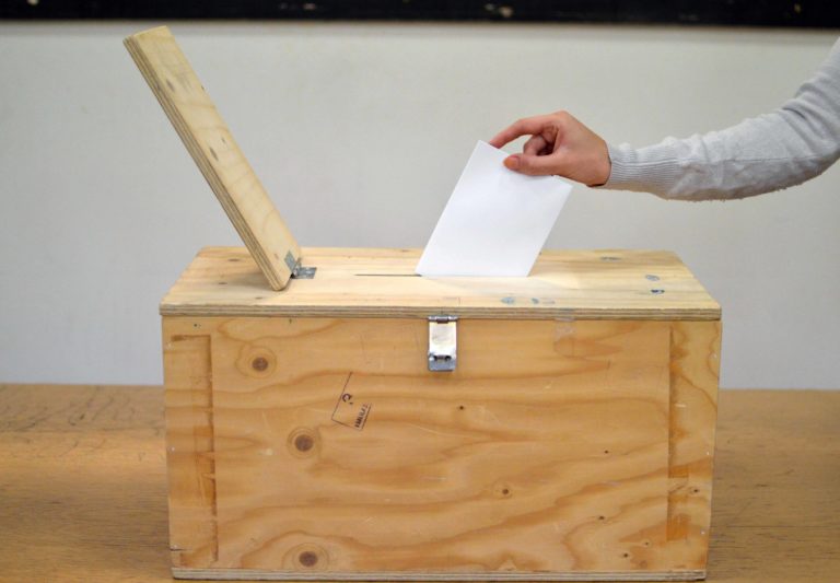 Vaaleissa äänestäjän käsi pudottaa vaalilipuketta puiseen vaalilippaaseen. Taustalla vaalea seinä.