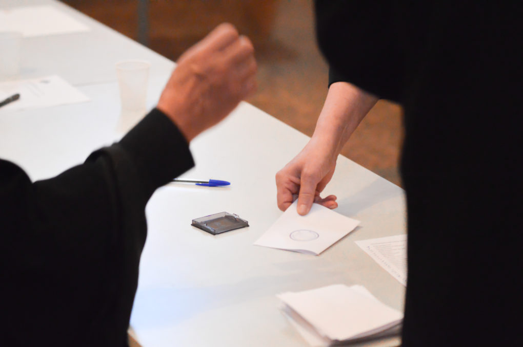 Äänestäjän käsi ojentaa äänestyslipukettaan vaalivirkailijan leimattavaksi vaalean pöydän päällä.