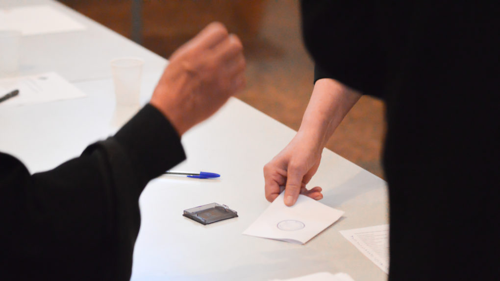 Äänestäjän käsi ojentaa äänestyslipukettaan vaalivirkailijan leimattavaksi vaalean pöydän päällä.