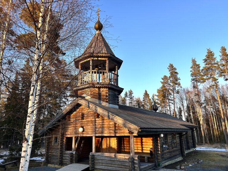 Kaunisniemen leirikeskuksen puinen kirkko, vieressä lehdettömiä koivuja. Aurinko valaisee kirkon etuseinää ja ovea kauniisti.