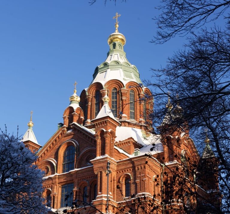 Uspenskin katedraali, katolla hieman lunta. Puiden oksat koristavat kuvan reunoja. Puiden oksat paljaat. Kupolin päällä hieman lunta. Taustalla sininen taivas.