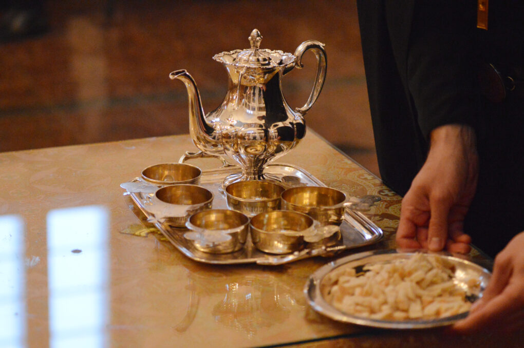 Pöydälä hopeinen kannu hopeisella tarjottimella ja siinä useita tyhjiä kuppejä, joita käytetään jälkiviinin tarjoamiseen. Vieressä antodora eli kirkkoleipä-lautanen, josta juuri ehtoollisella käynyt ottaa leipää. Kuvassa näkyy vain henkilön käsi ja musta hiha.