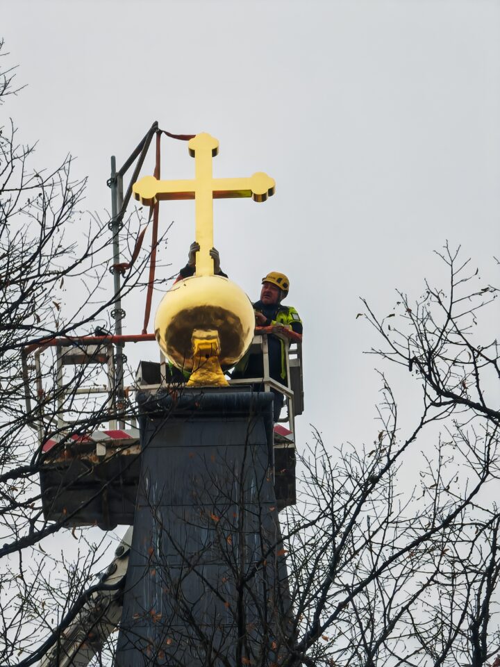 Kirkon kaolla lähikuvassa kullattu risti nostettuna paikoilleen ja ristin vieressä työmies telemässä viimeistetytöitä nosturista käsin. 
